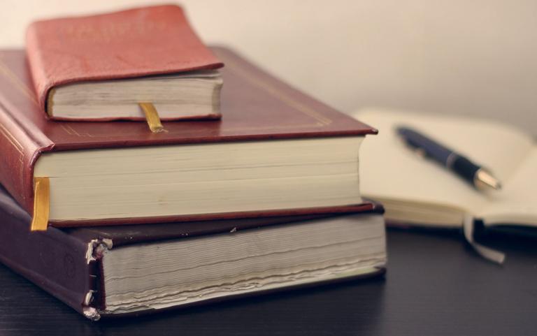 A stack of three closed books with bookmarks on a dark surface, accompanied by an open notebook and a pen.